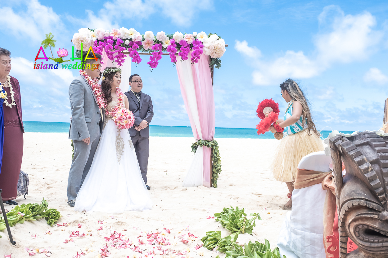 Bride, groom and father watching the hula dancer under their bamboo wedding arch on the beach at Waimanalo