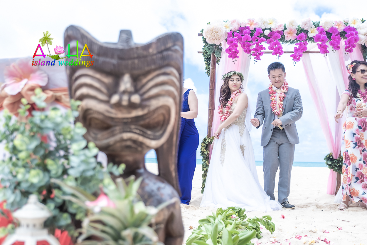 Face of a carved Hawaiian tiki with the bride learning the hula behind it