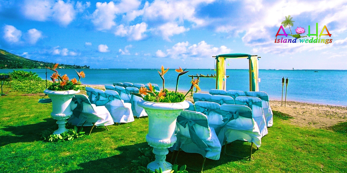Ceremony setup with white chairs, blue sashes and floral urns on a lawn by the ocean