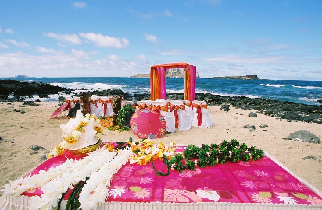 Ceremony table with white flower leis and a pink cloth, with the arch and the ocean behind