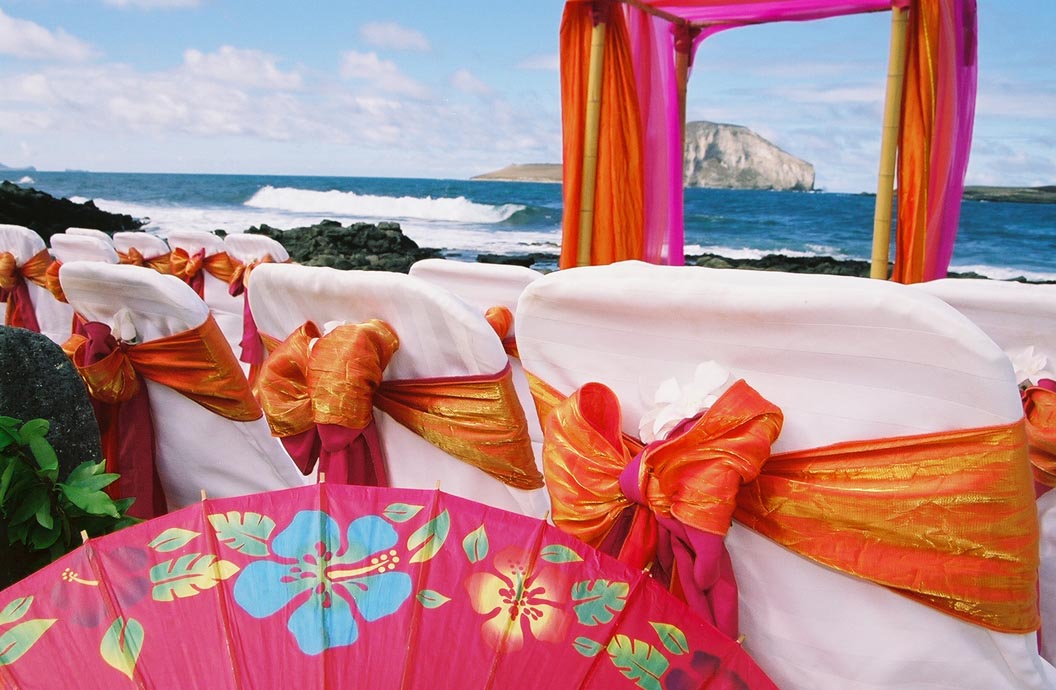 Close up of orange and fuchsia sashes on white chair covers beside the arch