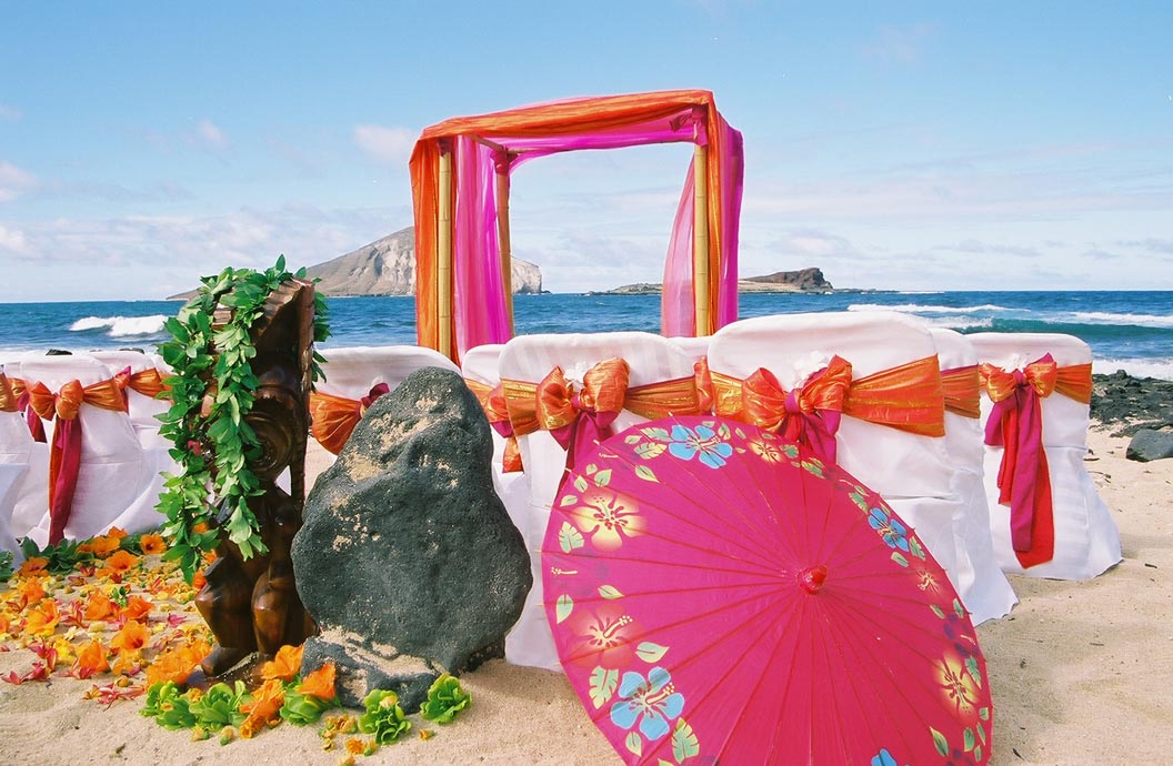 Orange and fuchsia bamboo arch by the ocean, with a lava rock wrapped in maile and a flowered pink parasol