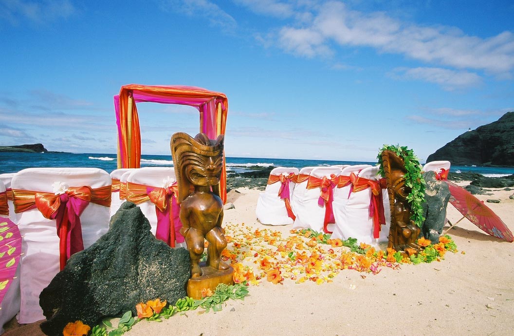 Two carved tikis by the orange and fuchsia arch, with orange plumerias scattered on the sand