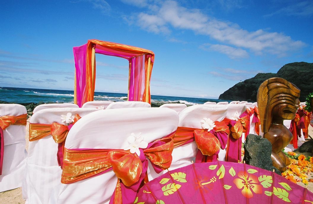 White chairs with orange and fuchsia sashes facing the arch, with a carved tiki and ocean view