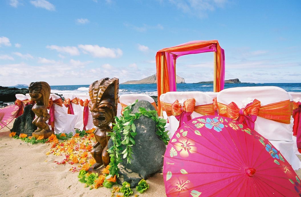 Tikis, a maile wrapped lava rock and a pink parasol beside the chairs and arch by the sea