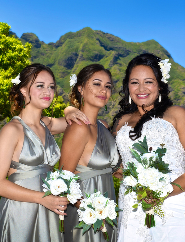 Bride and her bridesmaids in sage satin dresses holding white bouquets below Kualoa's mountains