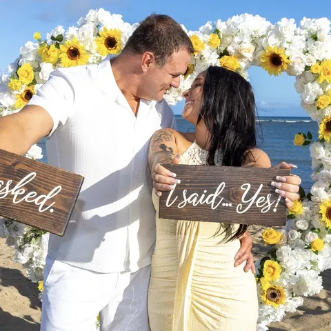 Engaged couple holding He asked and I said yes signs in the sunflower heart arch