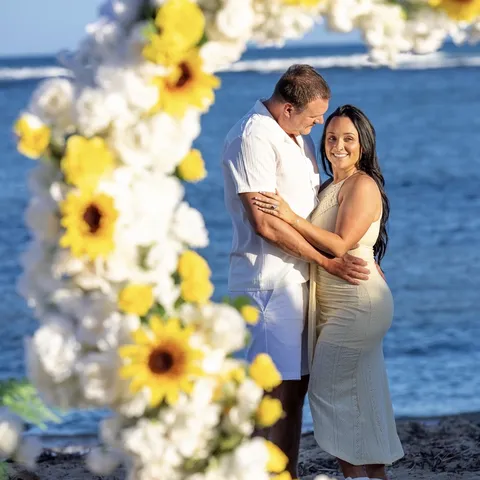 Couple hugging by the ocean, framed by the sunflower heart arch