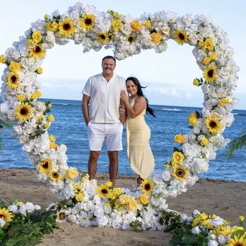 Makenzi and her fiancé under a heart arch of sunflowers and white flowers on an Oahu beach