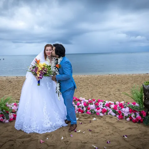 Sasha and Carlos kissing by a pink flower aisle and tiki statues on Kahala beach
