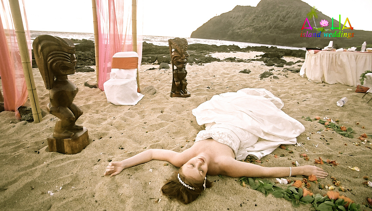 Bride lying on the sand between two carved tikis after her wedding