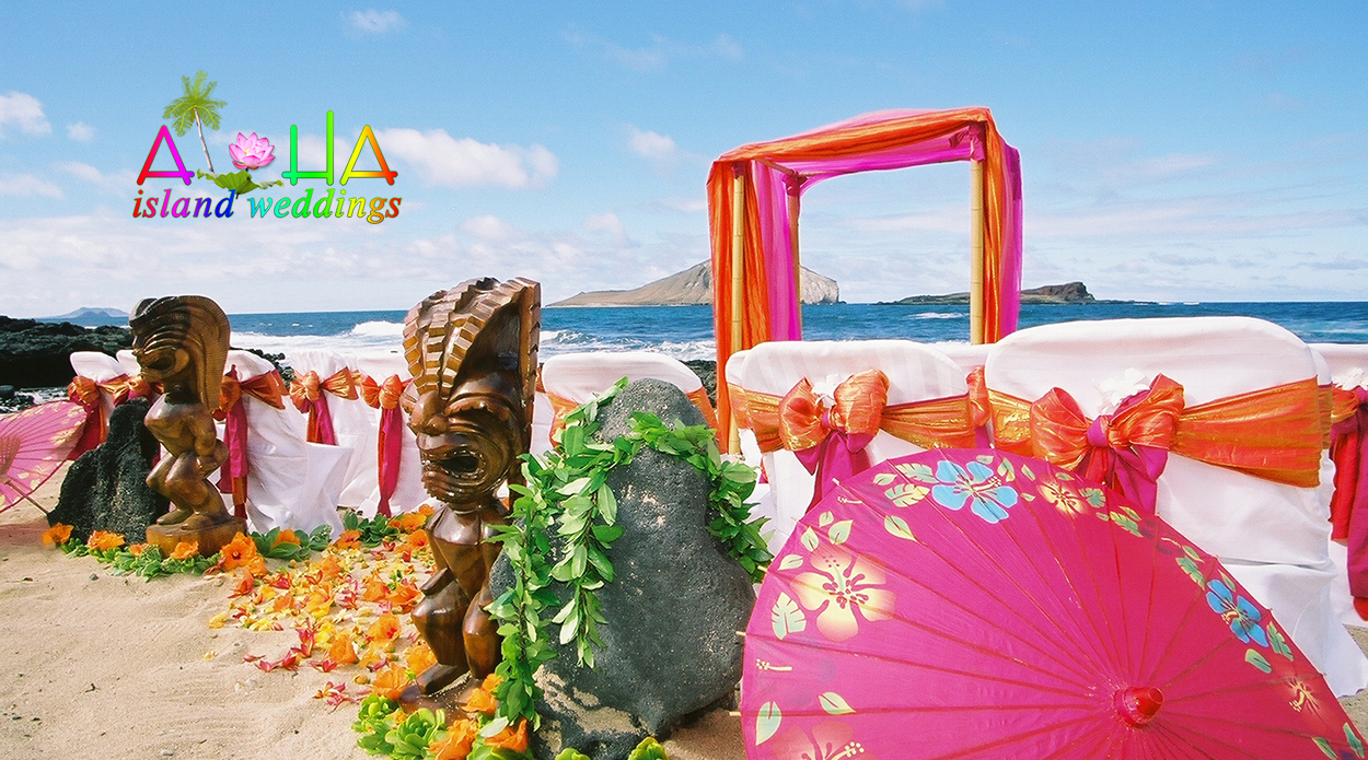 Carved tiki statues and a pink parasol beside an orange and fuchsia arch with white chairs on the beach