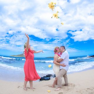 Daughter Emma tosses large yellow hibiscus in the air and her mom and dad kiss on the beach at makapuu